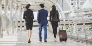 Three businesspeople, two women and one man, walk together along a modern, covered walkway. One woman is pulling a rolling suitcase. All are dressed in professional attire and walking away from the camera.