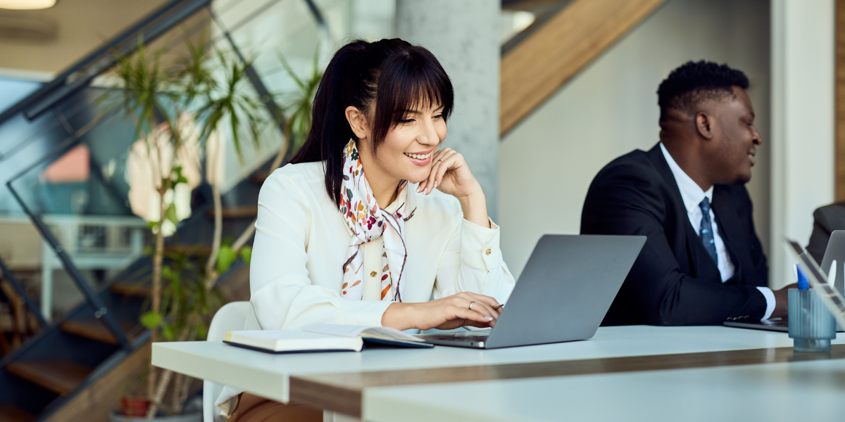 A woman smiling while working on a laptop at a desk in a modern office, with a notebook beside her. A man in a suit sits nearby, also working. Stairs and plants are visible in the background.