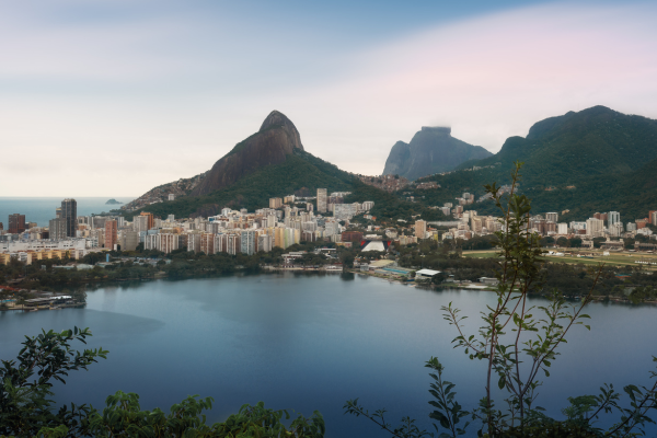 A panoramic view of Rio de Janeiro, Brazil, showing a calm lake in the foreground, city buildings, lush green mountains, and the iconic Sugarloaf Mountain under a cloudy sky.