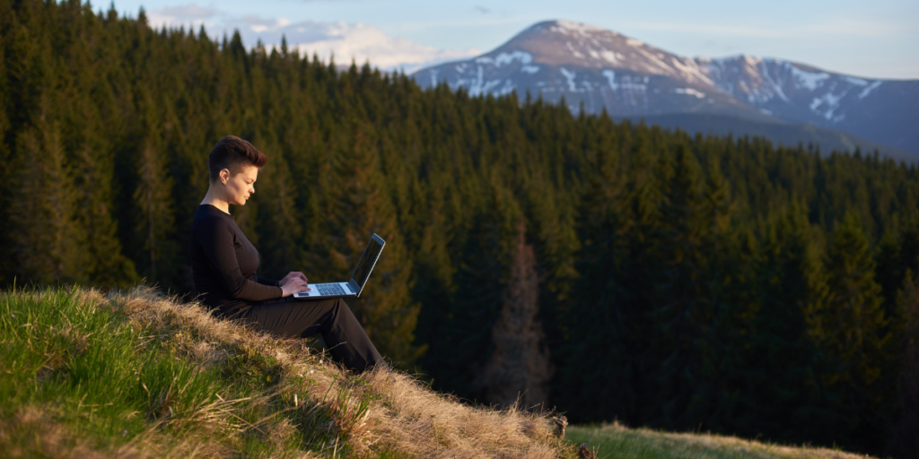 A person sits on a grassy hillside, working on a laptop, surrounded by dense pine forest with snowy mountains in the background under a clear sky.