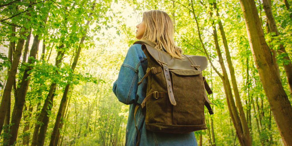 A person with long hair wearing a denim jacket and a brown backpack stands in a sunlit forest, looking up at the tall green trees around them.