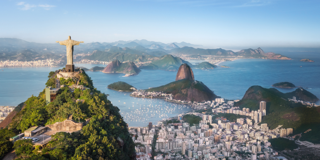 Aerial view of the Christ the Redeemer statue atop Corcovado Mountain overlooking Rio de Janeiro, with Sugarloaf Mountain, Guanabara Bay, and city buildings surrounded by lush green hills and blue water under a clear sky.