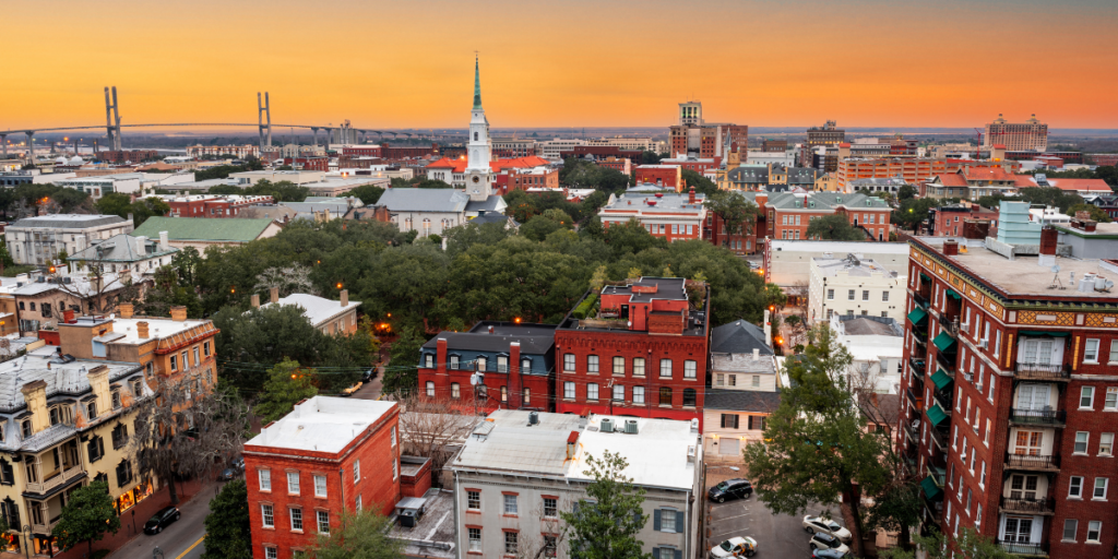 Aerial view of downtown Savannah, Georgia, at sunset, featuring historic buildings, tree-lined streets, a church steeple, and the Talmadge Memorial Bridge in the background.