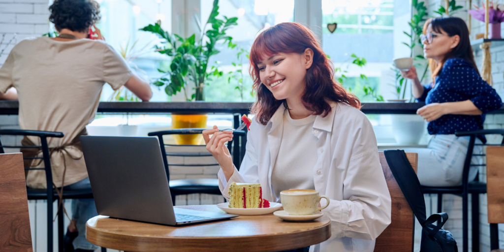 A young woman smiles while eating cake and using a laptop at a café. A cup of coffee sits on the table, and other people are visible in the background near large windows with plants.